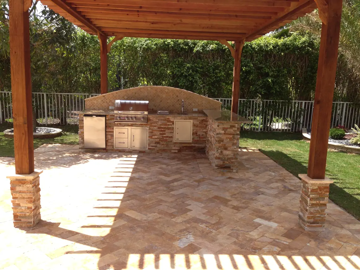 Stone outdoor kitchen with stainless steel grill beneath a wood pergola